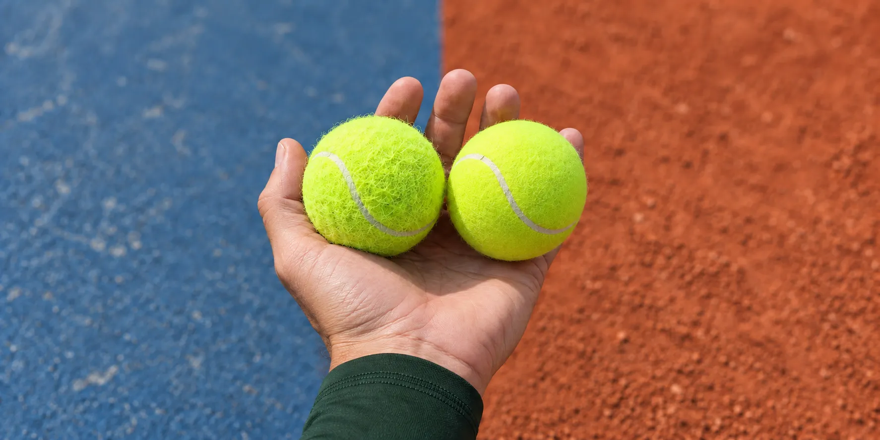 A hand holds two tennis balls above the boundary between a blue hard court and red clay