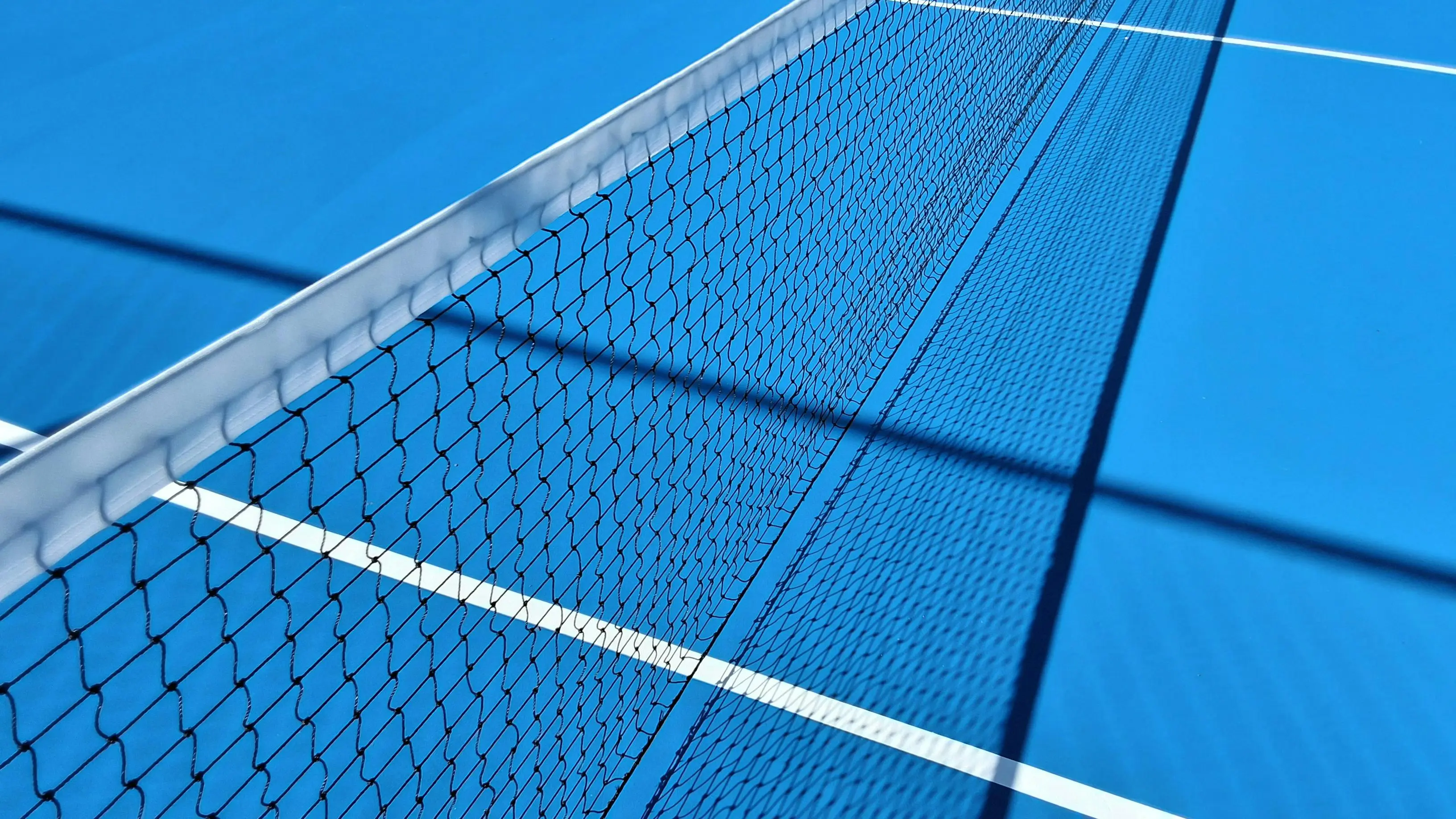 Primer plano de una pista dura de tenis azul con textura acrílica granular, línea blanca y pelota de tenis bajo luz cenital